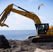 A yellow Caterpillar (CAT) 330 excavator is actively working on a coastal beach site, moving dark sediment or debris near the waterline. An operator is visible inside the cab, and a seagull is captured mid-flight above the machine. The background reveals a calm body of water under a partly cloudy blue sky.
