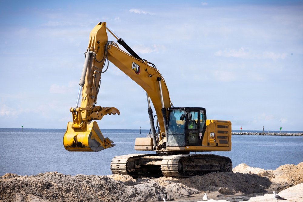 A yellow Caterpillar (CAT) excavator, model 330, is operating on a sandy coastal construction site near a body of water. The machine is equipped with a clamshell/grapple bucket attachment and appears to be engaged in dredging or shoreline construction work. A rock jetty or breakwater is visible in the background along with navigational buoys.