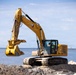 A yellow Caterpillar (CAT) excavator, model 330, is operating on a sandy coastal construction site near a body of water. The machine is equipped with a clamshell/grapple bucket attachment and appears to be engaged in dredging or shoreline construction work. A rock jetty or breakwater is visible in the background along with navigational buoys.