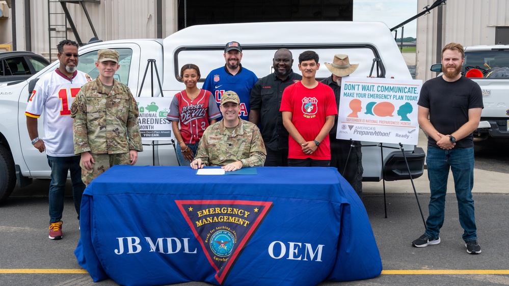 A group of approximately nine people pose for a photo at a Joint Base McGuire-Dix-Lakehurst (JB MDL) Office of Emergency Management (OEM) event for National Preparedness Month. A military officer in camouflage uniform is seated at a table covered with a blue tablecloth bearing the 'JB MDL OEM Emergency Management' insignia, appearing to sign a document. A sign in the background reads 'Don't Wait. Communicate. Make Your Emergency Plan Today' with FEMA and America's PrepareAthon branding.