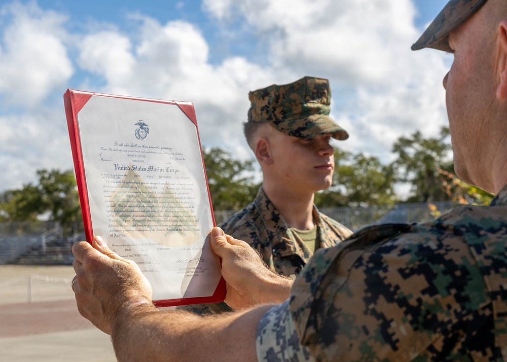 Two U.S. Marines in woodland MARPAT camouflage uniforms are participating in what appears to be a promotion or award ceremony outdoors. One Marine is holding up an official United States Marine Corps certificate in a red presentation folder, facing another Marine who is standing at attention. The document is clearly visible and bears the USMC eagle, globe, and anchor emblem.