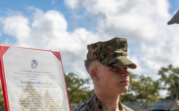 Two U.S. Marines in woodland MARPAT camouflage uniforms are participating in what appears to be a promotion or award ceremony outdoors. One Marine is holding up an official United States Marine Corps certificate in a red presentation folder, facing another Marine who is standing at attention. The document is clearly visible and bears the USMC eagle, globe, and anchor emblem.