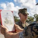 Two U.S. Marines in woodland MARPAT camouflage uniforms are participating in what appears to be a promotion or award ceremony outdoors. One Marine is holding up an official United States Marine Corps certificate in a red presentation folder, facing another Marine who is standing at attention. The document is clearly visible and bears the USMC eagle, globe, and anchor emblem.