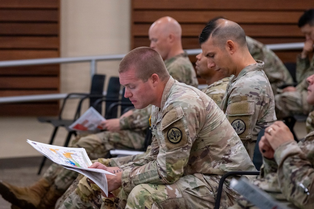 Multiple military personnel in U.S. Army Operational Camouflage Pattern (OCP) uniforms are seated in a briefing or training room, reviewing printed documents and materials. The foreground soldier is closely reading a colorful multi-page document, while others behind him are similarly engaged with their own materials. The setting appears to be an indoor auditorium or classroom environment.