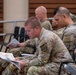 Multiple military personnel in U.S. Army Operational Camouflage Pattern (OCP) uniforms are seated in a briefing or training room, reviewing printed documents and materials. The foreground soldier is closely reading a colorful multi-page document, while others behind him are similarly engaged with their own materials. The setting appears to be an indoor auditorium or classroom environment.