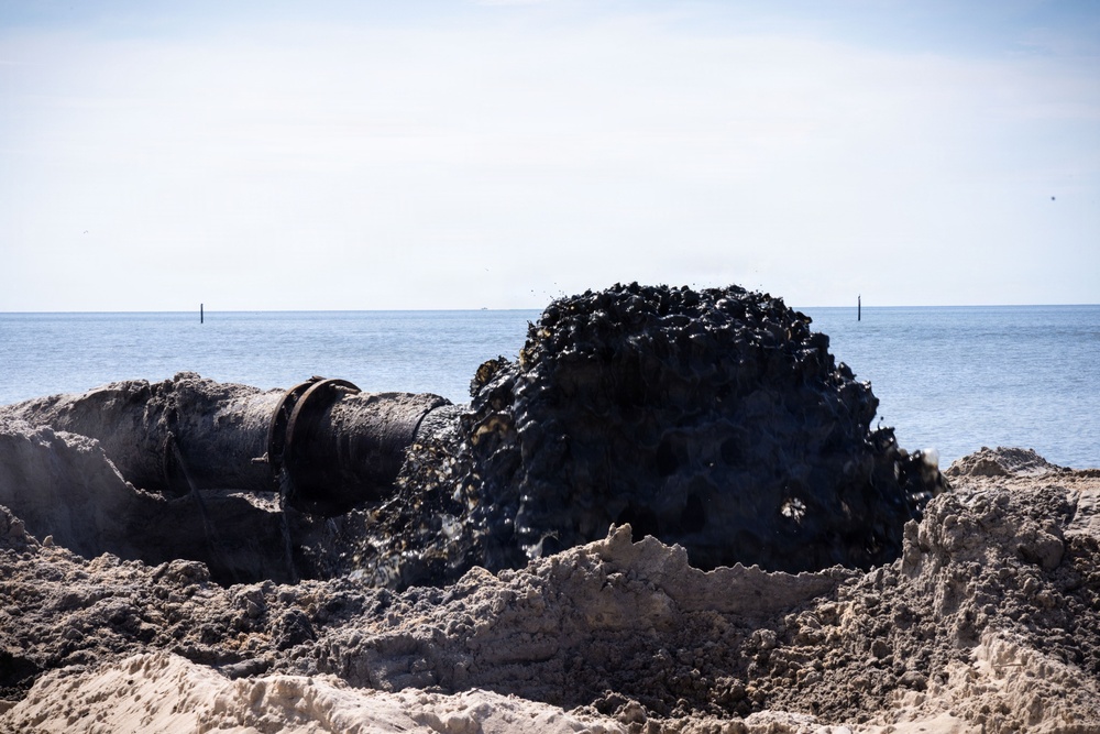 A large industrial pipe on a sandy beach is actively discharging a dark, viscous slurry or sediment mixture onto the shore. The material being expelled appears to be dark mud, silt, or possibly contaminated dredge material, forming a mound near the pipe's opening. The ocean is visible in the background under a clear sky, suggesting this may be a beach nourishment or dredging operation.