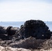A large industrial pipe on a sandy beach is actively discharging a dark, viscous slurry or sediment mixture onto the shore. The material being expelled appears to be dark mud, silt, or possibly contaminated dredge material, forming a mound near the pipe's opening. The ocean is visible in the background under a clear sky, suggesting this may be a beach nourishment or dredging operation.