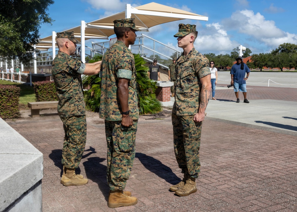 Three U.S. Marines in MARPAT woodland camouflage uniforms are engaged in what appears to be a formal military ceremony or inspection on a brick plaza. One Marine on the left is pinning or adjusting something on the sleeve/shoulder of the center Marine, while a third Marine stands at attention to the right. The setting appears to be a military base, likely a promotion or award ceremony.
