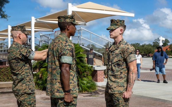 Three U.S. Marines in MARPAT woodland camouflage uniforms are engaged in what appears to be a formal military ceremony or inspection on a brick plaza. One Marine on the left is pinning or adjusting something on the sleeve/shoulder of the center Marine, while a third Marine stands at attention to the right. The setting appears to be a military base, likely a promotion or award ceremony.