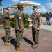 Three U.S. Marines in MARPAT woodland camouflage uniforms are engaged in what appears to be a formal military ceremony or inspection on a brick plaza. One Marine on the left is pinning or adjusting something on the sleeve/shoulder of the center Marine, while a third Marine stands at attention to the right. The setting appears to be a military base, likely a promotion or award ceremony.