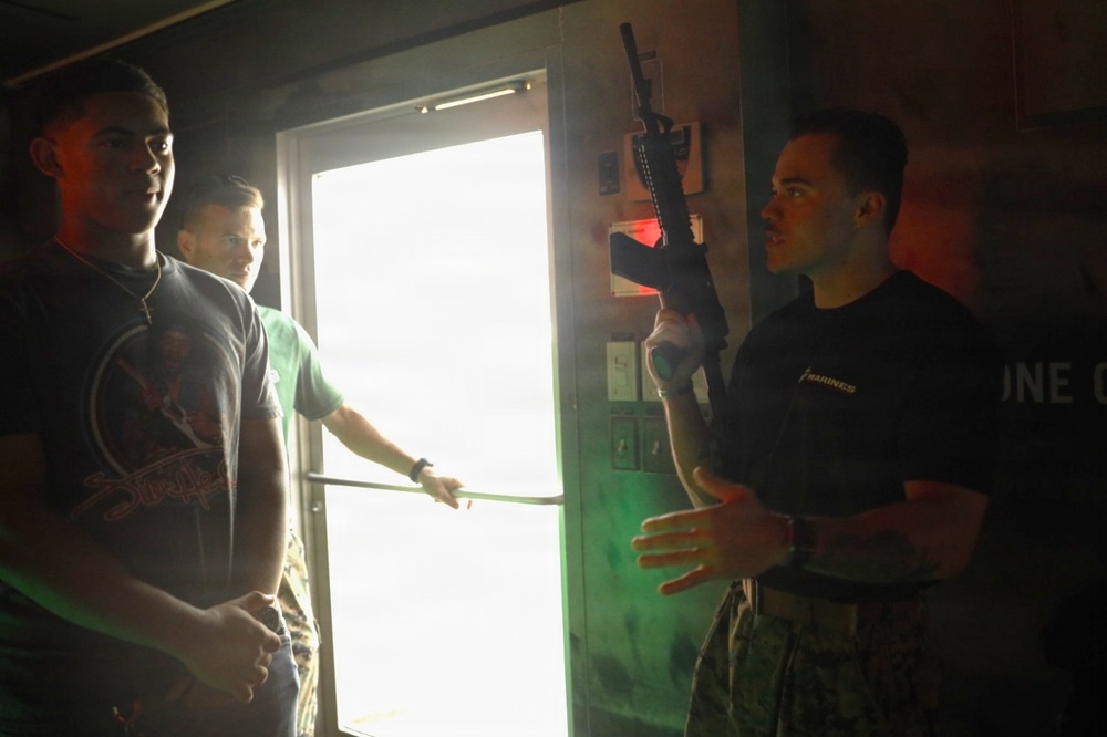 A person wearing a 'Marines' branded black t-shirt is holding up a rifle while appearing to explain or demonstrate something to two civilians in a dimly lit room. The setting appears to be a military recruitment or demonstration event, with the Marine instructor gesturing with his free hand. Two younger individuals in casual clothing are observing and listening.