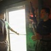 A person wearing a 'Marines' branded black t-shirt is holding up a rifle while appearing to explain or demonstrate something to two civilians in a dimly lit room. The setting appears to be a military recruitment or demonstration event, with the Marine instructor gesturing with his free hand. Two younger individuals in casual clothing are observing and listening.