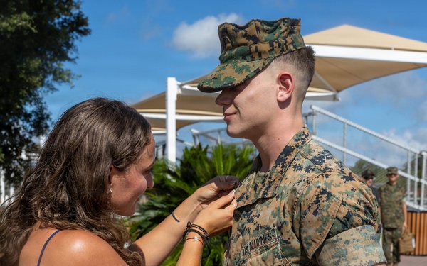 A woman in a blue dress is pinning something onto the uniform of a U.S. Marine in digital camouflage (MARPAT) during what appears to be a promotion or award ceremony. The Marine is standing at attention while the woman, likely a family member, performs the pinning. The setting appears to be an outdoor military installation on a sunny day.