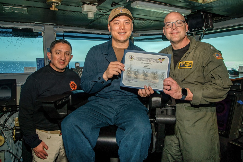 Three individuals are posing for a photo aboard what appears to be a U.S. Navy vessel's bridge or control room. The central figure, seated and wearing a blue coverall uniform and cap, is holding a certificate that reads 'USS Carl Vinson (CVN-70) Golden Eagle of the Day' dated 25 August 2020. The occasion appears to be an award or recognition ceremony.