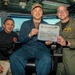 Three individuals are posing for a photo aboard what appears to be a U.S. Navy vessel's bridge or control room. The central figure, seated and wearing a blue coverall uniform and cap, is holding a certificate that reads 'USS Carl Vinson (CVN-70) Golden Eagle of the Day' dated 25 August 2020. The occasion appears to be an award or recognition ceremony.