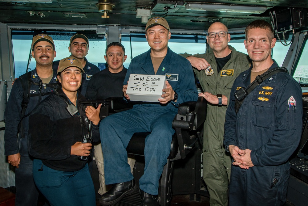 A group of U.S. Navy personnel pose for a photo on what appears to be the bridge of a naval vessel. The central figure is seated and holding a sign reading 'Gold Eagle of The Day,' suggesting a recognition or award ceremony. The group includes personnel in various Navy uniforms including flight suits and working uniforms.