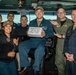 A group of U.S. Navy personnel pose for a photo on what appears to be the bridge of a naval vessel. The central figure is seated and holding a sign reading 'Gold Eagle of The Day,' suggesting a recognition or award ceremony. The group includes personnel in various Navy uniforms including flight suits and working uniforms.