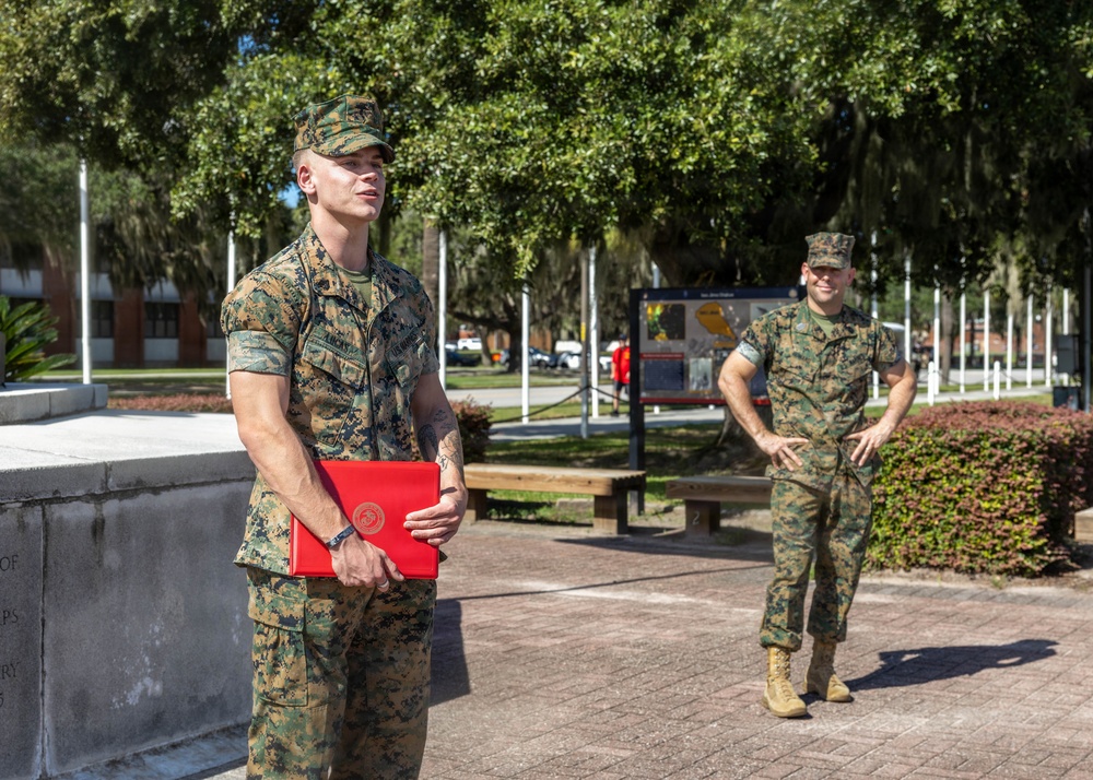 Two U.S. Marines in MARPAT woodland camouflage uniforms are standing outdoors on what appears to be a military base. The Marine in the foreground is holding a red folder bearing the U.S. Marine Corps emblem, suggesting a formal ceremony or award presentation has taken place or is in progress. The setting includes a monument, flagpoles, manicured landscaping, and large oak trees draped with Spanish moss.