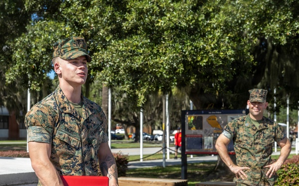 Two U.S. Marines in MARPAT woodland camouflage uniforms are standing outdoors on what appears to be a military base. The Marine in the foreground is holding a red folder bearing the U.S. Marine Corps emblem, suggesting a formal ceremony or award presentation has taken place or is in progress. The setting includes a monument, flagpoles, manicured landscaping, and large oak trees draped with Spanish moss.