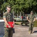 Two U.S. Marines in MARPAT woodland camouflage uniforms are standing outdoors on what appears to be a military base. The Marine in the foreground is holding a red folder bearing the U.S. Marine Corps emblem, suggesting a formal ceremony or award presentation has taken place or is in progress. The setting includes a monument, flagpoles, manicured landscaping, and large oak trees draped with Spanish moss.