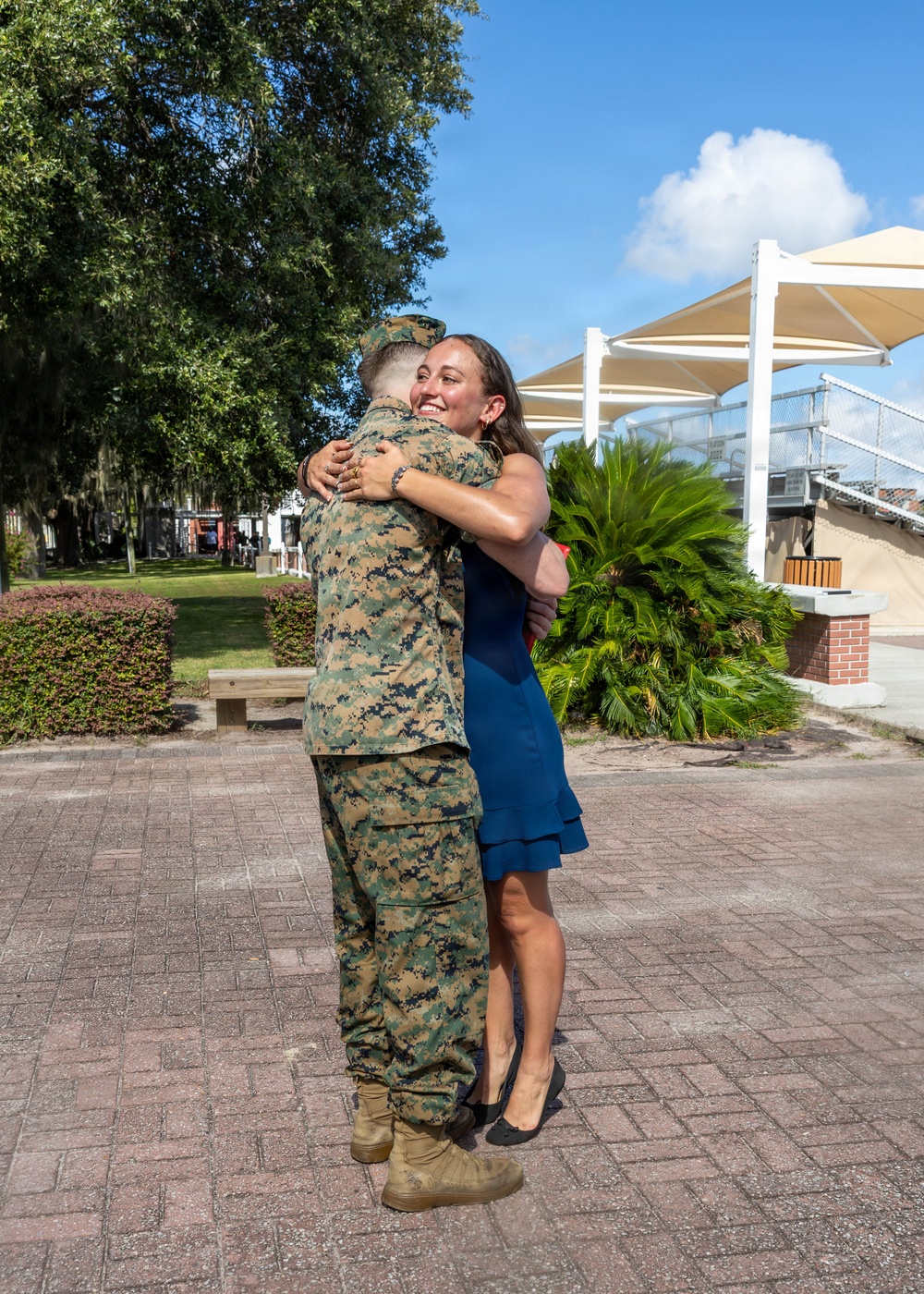 A person in U.S. Marine Corps MARPAT digital camouflage uniform is embracing a woman in a blue dress on a brick plaza. The woman is smiling broadly, suggesting an emotional reunion or celebratory moment. The setting appears to be a military base or institutional outdoor area with manicured landscaping and shade structures visible in the background.