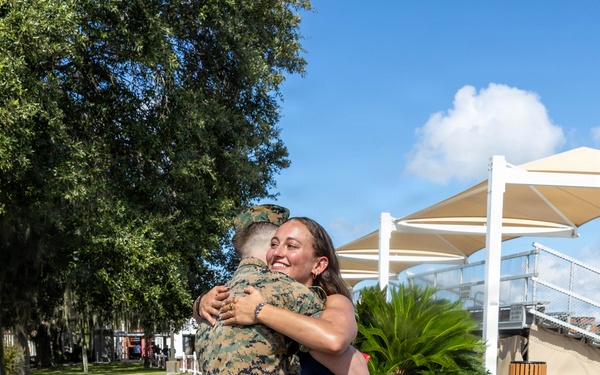 A person in U.S. Marine Corps MARPAT digital camouflage uniform is embracing a woman in a blue dress on a brick plaza. The woman is smiling broadly, suggesting an emotional reunion or celebratory moment. The setting appears to be a military base or institutional outdoor area with manicured landscaping and shade structures visible in the background.