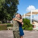 A person in U.S. Marine Corps MARPAT digital camouflage uniform is embracing a woman in a blue dress on a brick plaza. The woman is smiling broadly, suggesting an emotional reunion or celebratory moment. The setting appears to be a military base or institutional outdoor area with manicured landscaping and shade structures visible in the background.