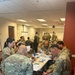 A group of U.S. Army soldiers in OCP (Operational Camouflage Pattern) uniforms are gathered in a classroom or conference room setting, engaged in what appears to be a training or briefing session. Multiple small groups are seated at tables reviewing printed materials and documents. The background shows additional soldiers gathered near whiteboards and flip charts.