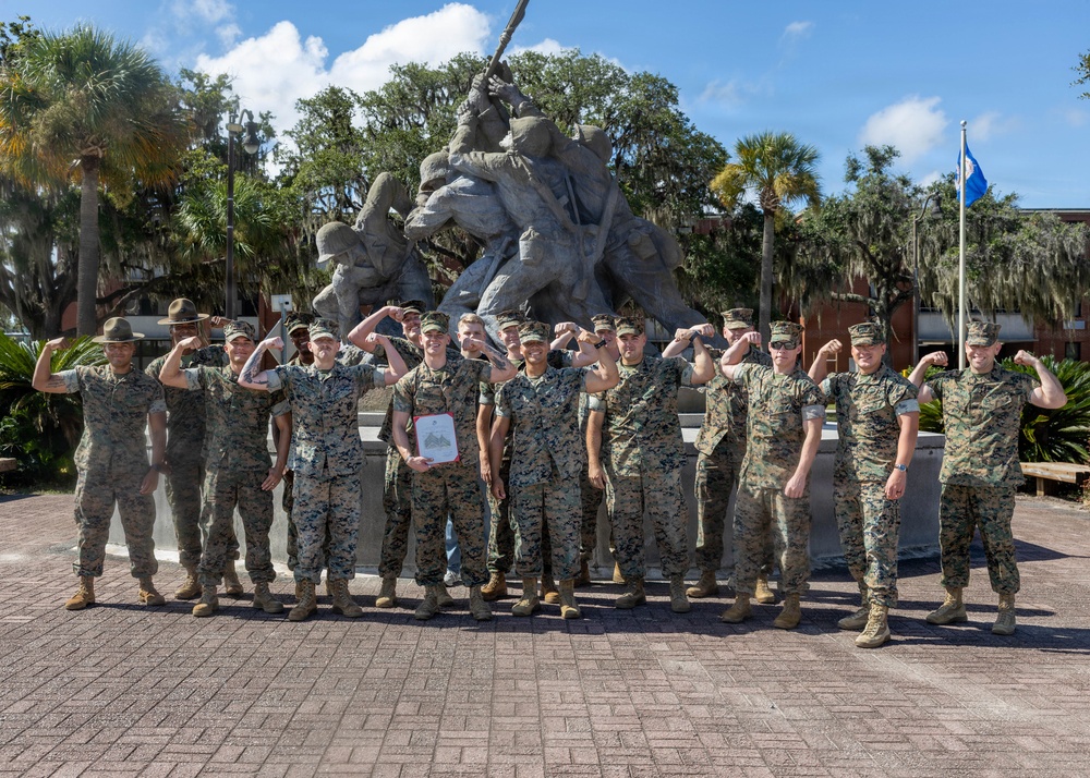 A group of approximately 15-16 military personnel in MARPAT camouflage uniforms pose for a group photo in front of a replica of the Iwo Jima flag-raising monument. Most individuals are flexing their arms in a celebratory pose, and one person in the center is holding a framed document or certificate. The setting appears to be a military installation, likely Marine Corps Recruit Depot (MCRD) Parris Island or a similar Marine Corps base, based on the monument and surroundings.