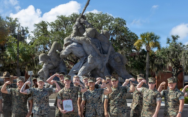 A group of approximately 15-16 military personnel in MARPAT camouflage uniforms pose for a group photo in front of a replica of the Iwo Jima flag-raising monument. Most individuals are flexing their arms in a celebratory pose, and one person in the center is holding a framed document or certificate. The setting appears to be a military installation, likely Marine Corps Recruit Depot (MCRD) Parris Island or a similar Marine Corps base, based on the monument and surroundings.