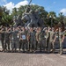 A group of approximately 15-16 military personnel in MARPAT camouflage uniforms pose for a group photo in front of a replica of the Iwo Jima flag-raising monument. Most individuals are flexing their arms in a celebratory pose, and one person in the center is holding a framed document or certificate. The setting appears to be a military installation, likely Marine Corps Recruit Depot (MCRD) Parris Island or a similar Marine Corps base, based on the monument and surroundings.