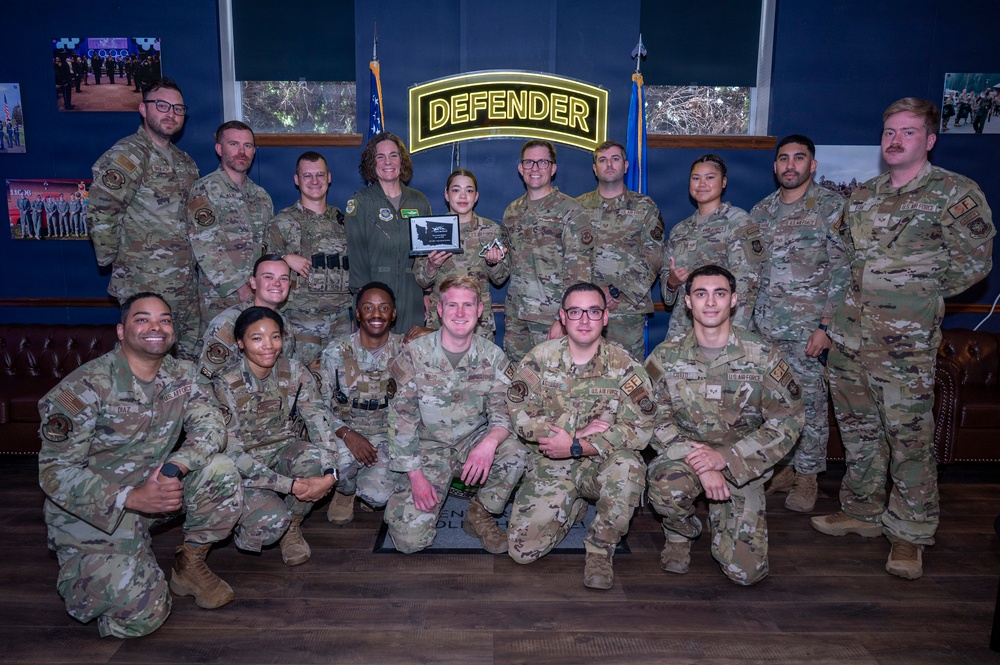 A group of approximately 18 U.S. Air Force military personnel pose for a formal group photo in front of an illuminated 'DEFENDER' sign. One individual in the center is holding a plaque or award, suggesting this is an award or recognition ceremony. The group is arranged in two rows with some members kneeling in front.