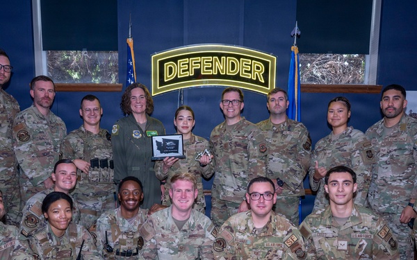 A group of approximately 18 U.S. Air Force military personnel pose for a formal group photo in front of an illuminated 'DEFENDER' sign. One individual in the center is holding a plaque or award, suggesting this is an award or recognition ceremony. The group is arranged in two rows with some members kneeling in front.