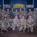 A group of approximately 18 U.S. Air Force military personnel pose for a formal group photo in front of an illuminated 'DEFENDER' sign. One individual in the center is holding a plaque or award, suggesting this is an award or recognition ceremony. The group is arranged in two rows with some members kneeling in front.