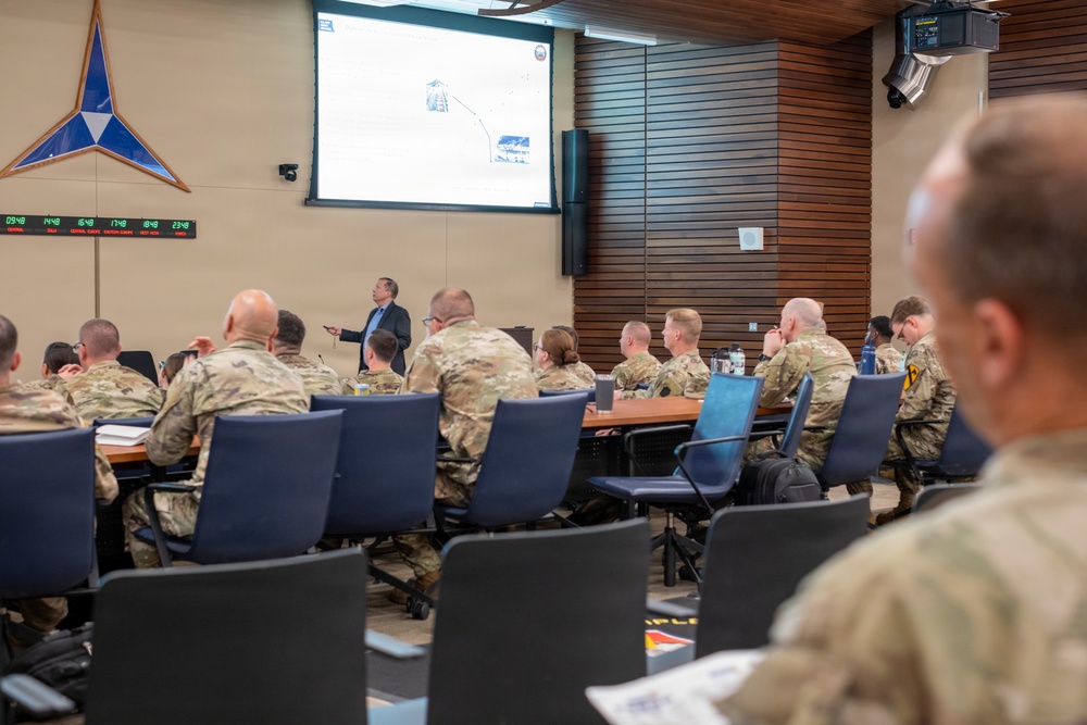 A civilian presenter is delivering a briefing or lecture to a room full of military personnel in camouflage uniforms. The setting appears to be a formal military conference or briefing room, equipped with a projection screen, multi-timezone digital clock display, and a NATO-style star emblem on the wall. Attendees are seated around conference tables facing the presenter and projected slide.