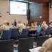 A civilian presenter is delivering a briefing or lecture to a room full of military personnel in camouflage uniforms. The setting appears to be a formal military conference or briefing room, equipped with a projection screen, multi-timezone digital clock display, and a NATO-style star emblem on the wall. Attendees are seated around conference tables facing the presenter and projected slide.
