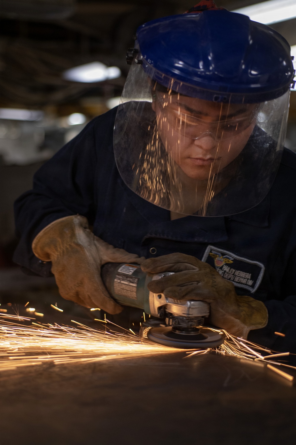 A worker wearing full protective gear — including a blue hard hat with clear face shield, safety glasses, and leather gloves — operates an angle grinder on a metal surface. Bright orange sparks are flying dramatically from the grinding point, illuminating the scene. A name patch is visible on the dark blue work uniform, partially readable as 'HERRERA' with additional text indicating a department or unit.
