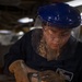 A worker wearing full protective gear — including a blue hard hat with clear face shield, safety glasses, and leather gloves — operates an angle grinder on a metal surface. Bright orange sparks are flying dramatically from the grinding point, illuminating the scene. A name patch is visible on the dark blue work uniform, partially readable as 'HERRERA' with additional text indicating a department or unit.
