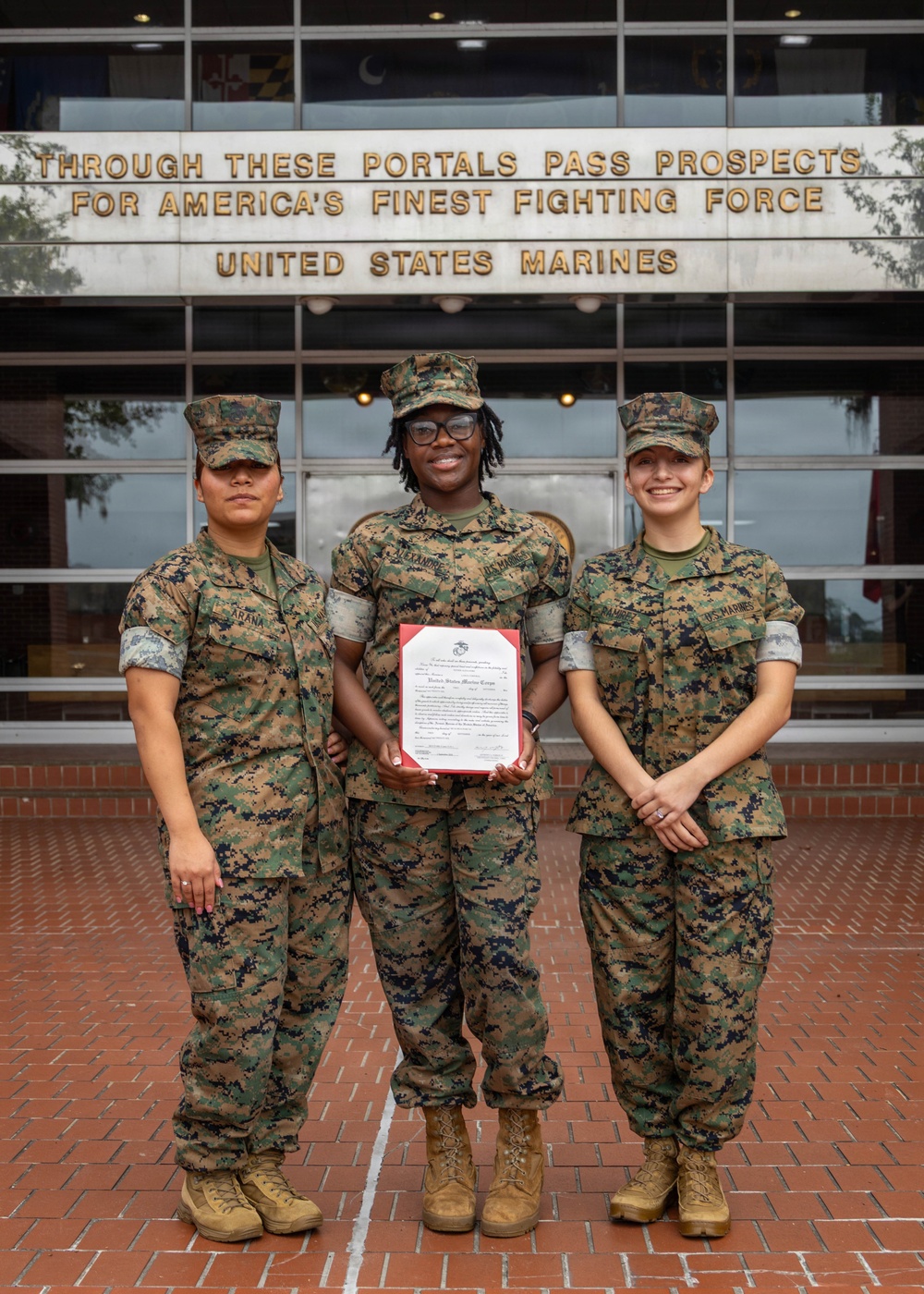 Three United States Marines in MARPAT camouflage uniforms pose for a photo in front of a building bearing the inscription 'Through These Portals Pass Prospects for America's Finest Fighting Force - United States Marines.' The center Marine is holding a framed certificate or official document, suggesting a recognition or award ceremony. The setting appears to be a Marine Corps recruiting or administrative facility.