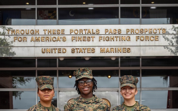 Three United States Marines in MARPAT camouflage uniforms pose for a photo in front of a building bearing the inscription 'Through These Portals Pass Prospects for America's Finest Fighting Force - United States Marines.' The center Marine is holding a framed certificate or official document, suggesting a recognition or award ceremony. The setting appears to be a Marine Corps recruiting or administrative facility.