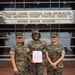 Three United States Marines in MARPAT camouflage uniforms pose for a photo in front of a building bearing the inscription 'Through These Portals Pass Prospects for America's Finest Fighting Force - United States Marines.' The center Marine is holding a framed certificate or official document, suggesting a recognition or award ceremony. The setting appears to be a Marine Corps recruiting or administrative facility.