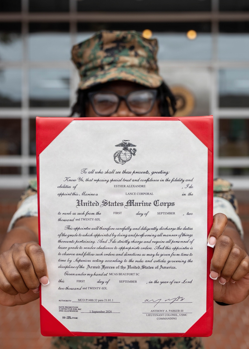 A United States Marine in camouflage uniform and cover holds up an official promotion certificate from the United States Marine Corps. The certificate appoints Esther Alexandre to the rank of Lance Corporal, effective 1 September 2026, signed by Lieutenant Colonel Anthony A. Parker III at MCAS Beaufort, SC. The document is displayed in a red presentation folder with the USMC Eagle, Globe, and Anchor emblem prominently visible.