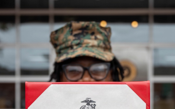 A United States Marine in camouflage uniform and cover holds up an official promotion certificate from the United States Marine Corps. The certificate appoints Esther Alexandre to the rank of Lance Corporal, effective 1 September 2026, signed by Lieutenant Colonel Anthony A. Parker III at MCAS Beaufort, SC. The document is displayed in a red presentation folder with the USMC Eagle, Globe, and Anchor emblem prominently visible.