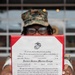 A United States Marine in camouflage uniform and cover holds up an official promotion certificate from the United States Marine Corps. The certificate appoints Esther Alexandre to the rank of Lance Corporal, effective 1 September 2026, signed by Lieutenant Colonel Anthony A. Parker III at MCAS Beaufort, SC. The document is displayed in a red presentation folder with the USMC Eagle, Globe, and Anchor emblem prominently visible.