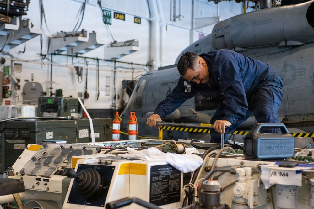 A U.S. Navy sailor in blue coveralls is performing maintenance work on helicopter equipment inside what appears to be a naval vessel's hangar bay. The technician is using a torque wrench or similar tool on an open mechanical assembly, with a military helicopter visible in the background. The workspace is cluttered with tools, equipment cases, and safety cones, indicating an active maintenance operation.