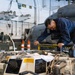 A U.S. Navy sailor in blue coveralls is performing maintenance work on helicopter equipment inside what appears to be a naval vessel's hangar bay. The technician is using a torque wrench or similar tool on an open mechanical assembly, with a military helicopter visible in the background. The workspace is cluttered with tools, equipment cases, and safety cones, indicating an active maintenance operation.
