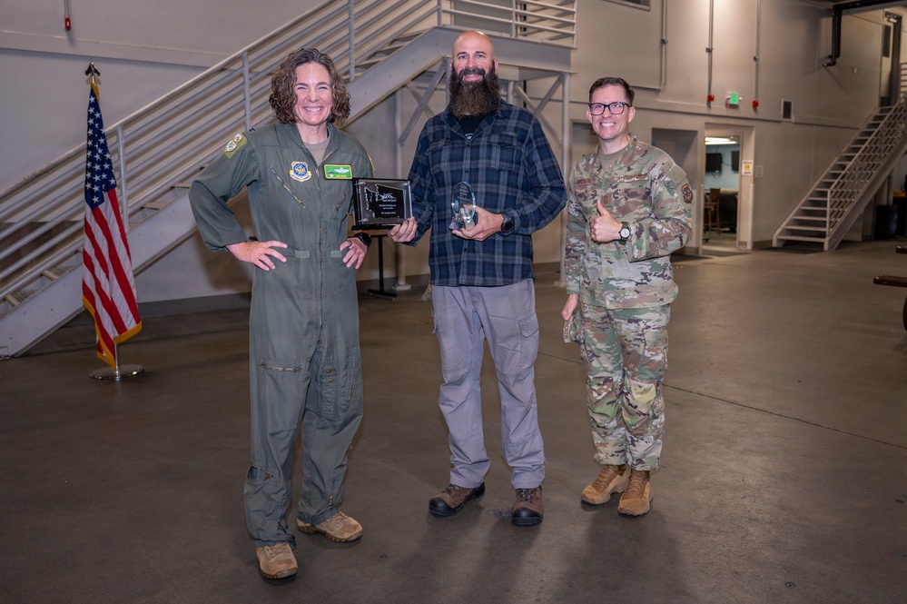 Three individuals pose for a photo in what appears to be a military hangar or large indoor facility. The central figure, a civilian male with a beard, holds two awards — a plaque and a crystal trophy — flanked by two military personnel in uniform. The occasion appears to be an award or recognition ceremony.