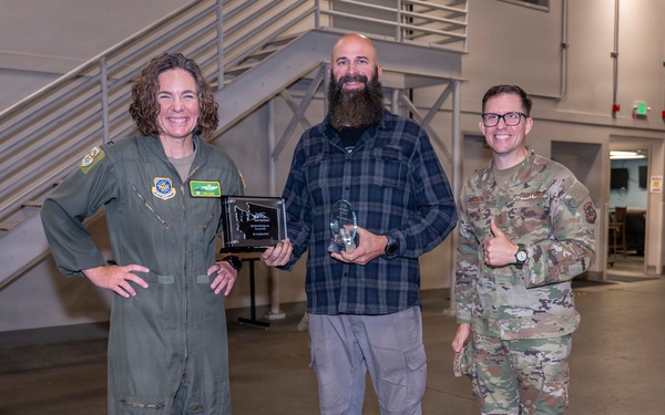Three individuals pose for a photo in what appears to be a military hangar or large indoor facility. The central figure, a civilian male with a beard, holds two awards — a plaque and a crystal trophy — flanked by two military personnel in uniform. The occasion appears to be an award or recognition ceremony.