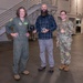 Three individuals pose for a photo in what appears to be a military hangar or large indoor facility. The central figure, a civilian male with a beard, holds two awards — a plaque and a crystal trophy — flanked by two military personnel in uniform. The occasion appears to be an award or recognition ceremony.