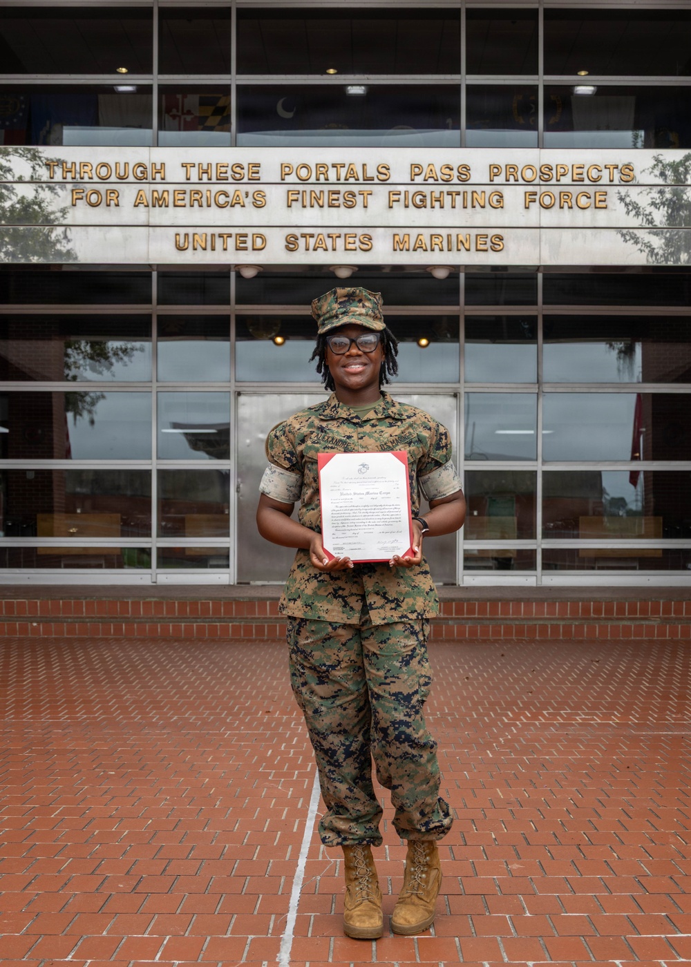 A United States Marine in woodland MARPAT camouflage uniform stands proudly in front of a building bearing the inscription 'Through These Portals Pass Prospects For America's Finest Fighting Force - United States Marines.' The Marine is holding an official document, likely a certificate or enlistment/promotion order with a red border, and is smiling at the camera. The setting appears to be a Marine Corps recruiting station or administrative building.